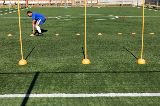 Male soccer player wearing blue jersey bending over picking orange cone on turf, copy space