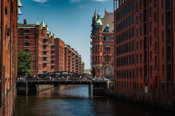 Naklejka premium Canal view through Hamburg’s Speicherstadt reveals pointed green rooftops, ornate facades, and multiple bridges connecting the red-brick buildings.