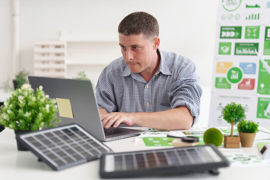 Adult Caucasian man working on laptop analyzing ESG data and green business strategies with desk solar panels for carbon footprint reduction