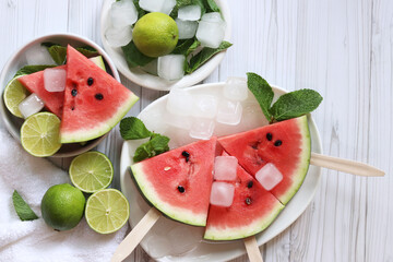Watermelon slices, limes and mint in white plates, top view. Ripe watermelon pieces on sticks, wooden table and lime halves. Summer food. Summer fruits background with ice. Flat lay. Products