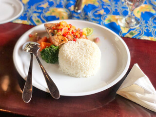 Delicious Indonesian dish featuring grilled salmon topped with spicy sambal dabu-dabu, served with steamed rice and vegetables on a white plate at a table.
