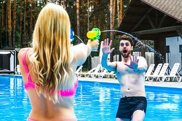 Pretty lady aiming at a man with a water gun while playing in the swimming pool outdoor. Summer vacation, active leisure, lifestyle concept