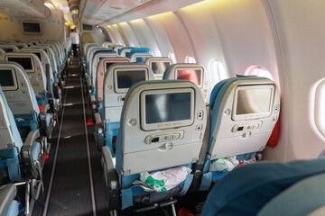 Rows of blue seats fill the airplane cabin, with overhead lights dimmed. Passengers prepare for departure, adding trash to nearby seat pockets as flight attendants pass through.
