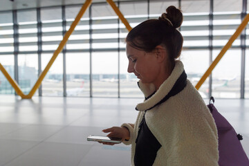 A woman stands in a busy airport terminal looking at her mobile phone. Natural light floods the space through large windows as planes are visible in the background.