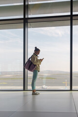 A traveler stands in the airport terminal, focused on her phone. Large windows provide a view of the runway and distant mountains under a clear sky.