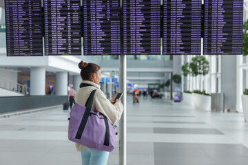 A woman stands at an airport terminal, focused on her phone while looking at the large flight information display board. The terminal is bustling with travelers in the background.