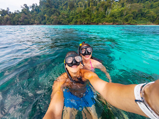 Two individuals enjoy snorkeling in clear tropical waters surrounded by lush greenery. They smile for a selfie while exploring the vibrant marine life together.