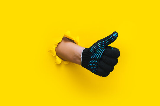 A left man's hand in a black fabric work glove shows a thumb up (like). Torn hole in yellow paper. Good job, fun mood and approval concept. Copy space. - Powered by Adobe