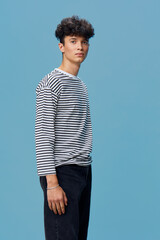 young man wearing striped long sleeve shirt and black jeans standing against blue background with...