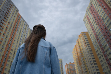 A young woman stands with her back to the camera looking up at tall, brightly colored apartment...