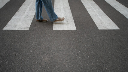 A person is crossing a pedestrian crosswalk with white stripes, wearing casual denim jeans and...