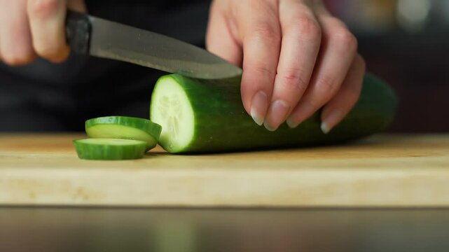Hands slicing a cucumber on a wooden cutting board with a knife, preparing food.