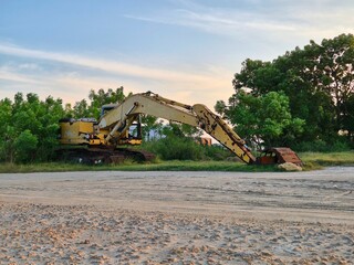 Old yellow excavator with rusted bucket parked near trees on a construction site at sunset sky