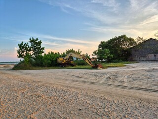 Old yellow excavator with rusted bucket parked near trees on a construction site at sunset sky