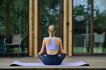 Back view of a woman sits on a yoga mat placed on a wooden terrace of tiny house