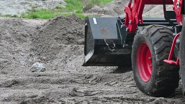 Close-up shot of a heavy-duty tractor loader scooping soil with a metal bucket at a construction site. The red front loader moves through dirt and gravel while performing earthmoving work.