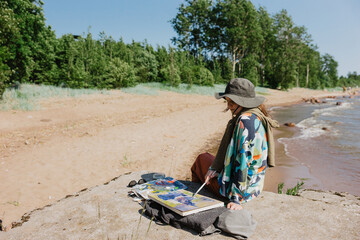 A young artist girl is sitting on the beach painting a sunny day with strong winds and waves on the...