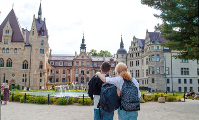 A man and a woman stand embracing on the square in front of the castle. Tourists admire the architecture of the old Moszna Castle in Poland