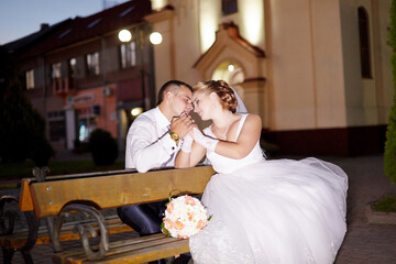 A bride and groom are sitting on a bench, holding hands and smiling