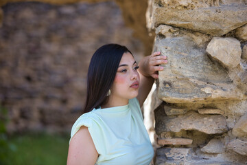 A beautiful Spanish teenager with long dark hair rests her hand on a stone wall and looks sad and...