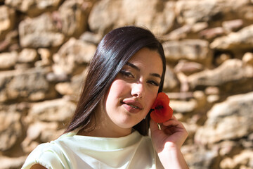 A beautiful Spanish teenager with long dark hair picks up a delicate, beautiful red poppy and holds it next to her face. The woman is happy and passionate.