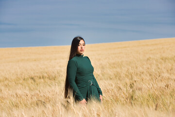 A beautiful, dark-haired teenage Latin girl dressed in green stands among a dry, yellow wheat field on a summer day. In the background, the blue sky.