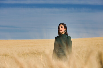 A beautiful, dark-haired teenage Latin girl dressed in green stands among a dry, yellow wheat field on a summer day. In the background, the blue sky.