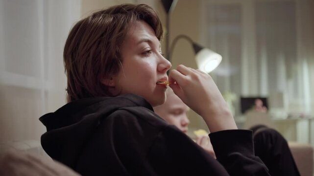 Young mother and her son wearing hearing aids share popcorn while laughing at a TV show. Warm living room scene showcasing joyful family bonding and inclusive representation. Focus on normalizing assi