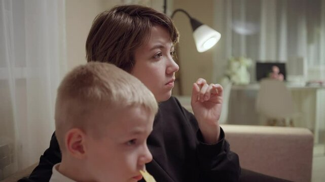 Young mother and her son wearing hearing aids share popcorn while laughing at a TV show. Warm living room scene showcasing joyful family bonding and inclusive representation. Focus on normalizing assi