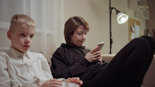 Young mother and her son wearing hearing aids share popcorn while laughing at a TV show. Warm living room scene showcasing joyful family bonding and inclusive representation. Focus on normalizing assi