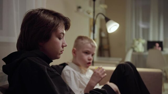 Young mother and her son wearing hearing aids share popcorn while laughing at a TV show. Warm living room scene showcasing joyful family bonding and inclusive representation. Focus on normalizing assi