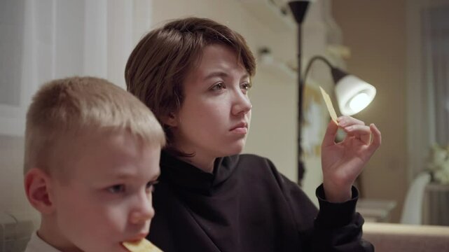 Young mother and her son wearing hearing aids share popcorn while laughing at a TV show. Warm living room scene showcasing joyful family bonding and inclusive representation. Focus on normalizing assi