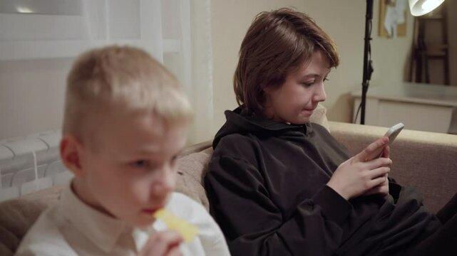 Young mother and her son wearing hearing aids share popcorn while laughing at a TV show. Warm living room scene showcasing joyful family bonding and inclusive representation. Focus on normalizing assi