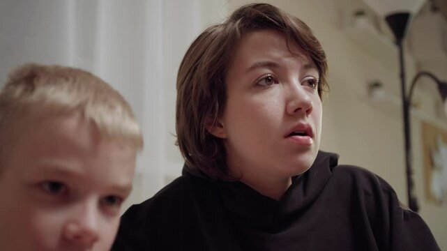 Young mother and her son wearing hearing aids share popcorn while laughing at a TV show. Warm living room scene showcasing joyful family bonding and inclusive representation. Focus on normalizing assi