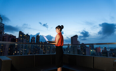 Fototapeta premium Young beautiful asian businesswoman with laptop on the building rooftop at city night