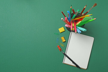 Top view of school supplies, pencils, erasers, paper clips and notebooks, on a green background....