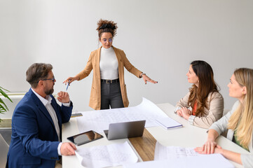 Confident female manager with blueprint on table explaining project details to colleagues in...