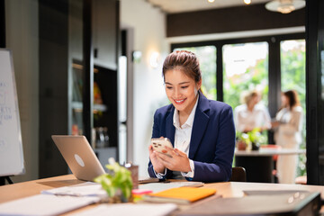 Young asian  Businesswoman  using  mobile phone reading message or  e mail  and working online on laptop web surfing information while sitting in modern office.