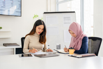 Two asian businesswomen are collaborating on a project, analyzing financial data and taking notes,...