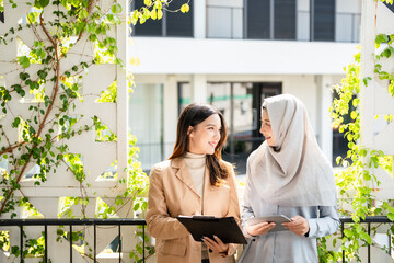 Asian business professional woman talking to Muslim female coworker in hijab, discussing...