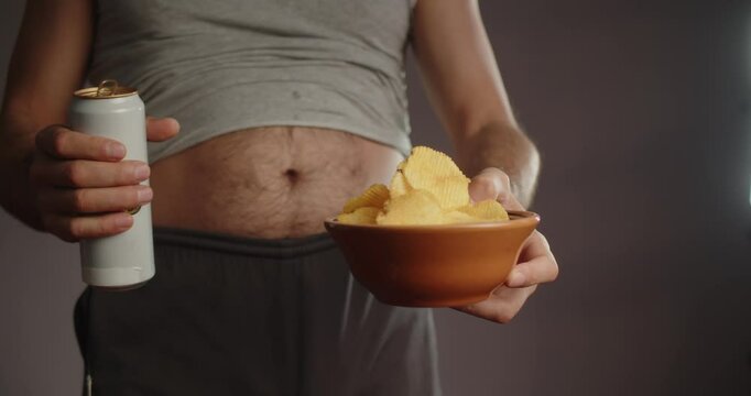 Modern Adult man in sweatpants and a T-shirt with a bare hairy beer belly offers a metal can of drink and chips. Close-up on the stomach and arms