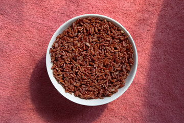 Top view of a white bowl filled with cooked brown rice on textured pink surface background.       
