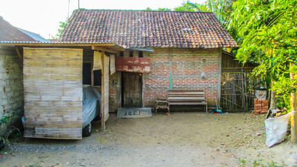 A traditional village house with brick walls, bamboo canopy, wooden benches, and a car covered by a protective sheet in a dirt yard.


