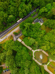A drone view of a road cutting through dense green forest, with open grassy fields on one side and...