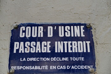 Cour d'usine,. Passage interdit. La direction décline toute responsabilité en cas d'accident.  Plaque émaillée bleue et blanche ancienne sur un mur blanc.