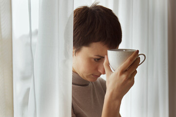 Sad woman with a cup stands near the window in her house.