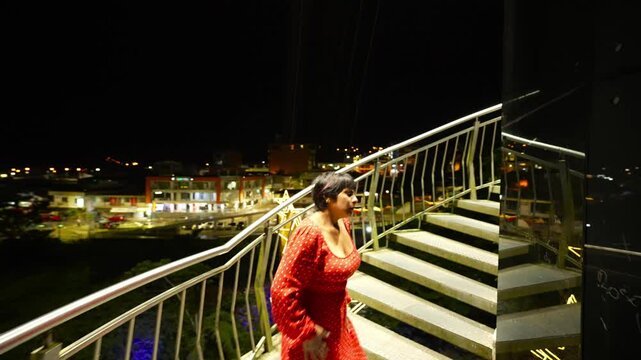Female tourist in elegant red dress ascending monumental stairs during nighttime urban exploration, illuminated by city lights with architectural backdrop in Tena, Ecuador