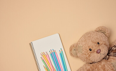 Wooden colored pencils and open notebook, brown teddy bear next to it on beige background