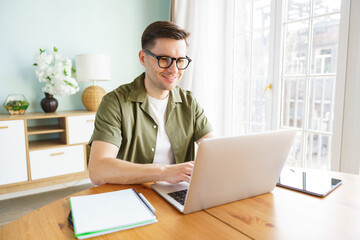 Young man enjoys working remotely at home, focusing on his laptop while surrounded by a bright and...