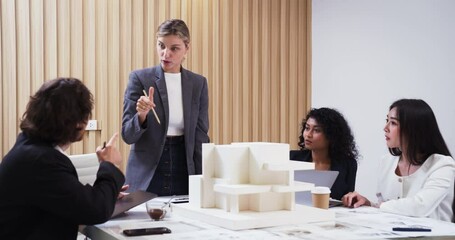 Confident female architect leads a collaborative design discussion in a bright, modern office. She stands beside a detailed architectural model, presenting ideas to a diverse team. - Powered by Adobe